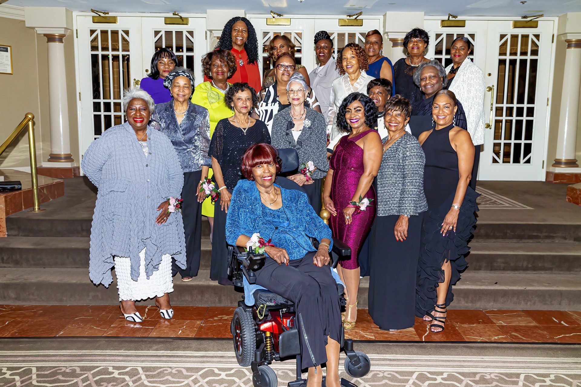 A group of diverse women, dressed elegantly, gather on steps for a celebration, showcasing unity and joy at a special event.